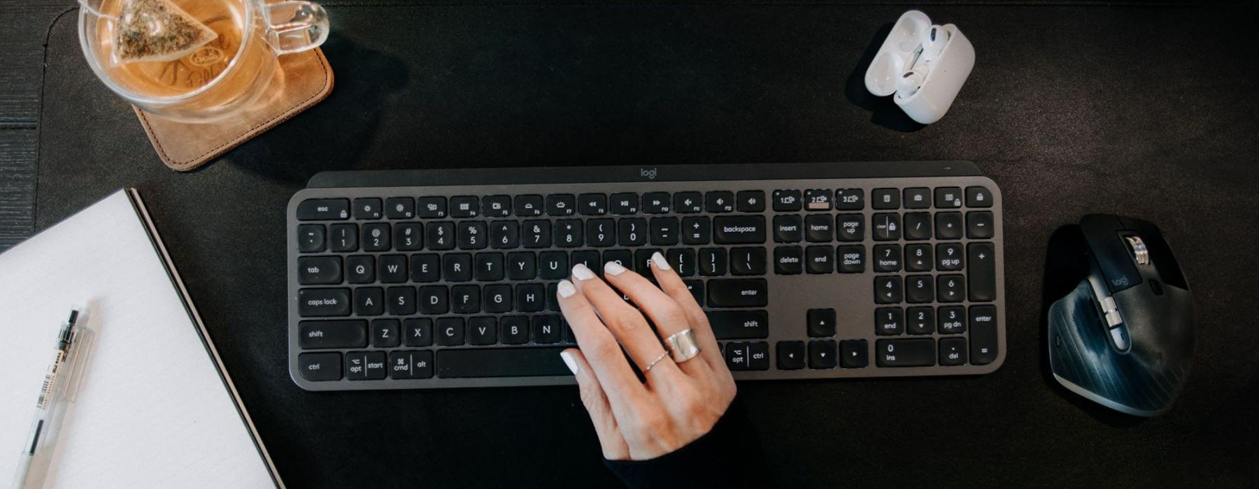 woman's hand on a keyboard surrounded by office items and a cup of tea on a coaster